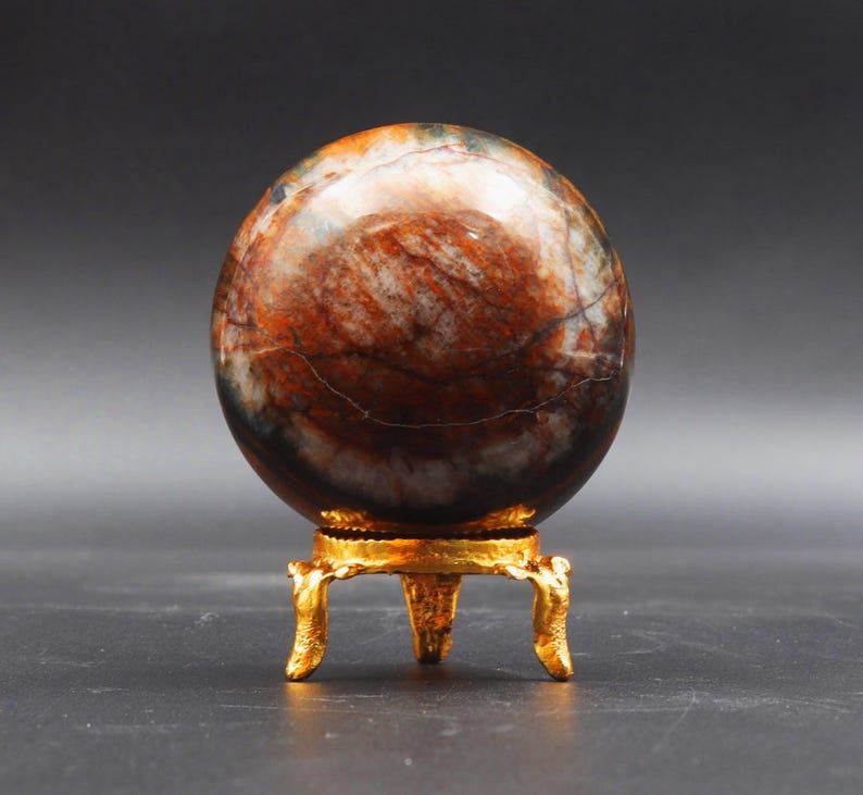 Puede incluir: Una esfera pulida de &aacute;gata multicolor sobre un soporte de metal dorado con tres patas. La esfera tiene un patr&oacute;n de m&aacute;rmol marr&oacute;n, blanco y negro.