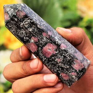May include: A polished, rectangular gemstone obelisk. The stone is primarily black with a speckled pattern of white and gray, and features several large, round, deep pink inclusions. The obelisk is held in a hand, with a blurred green and yellow background.