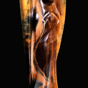 Peut inclure: Une figurine d'ange sculpt&eacute;e en pierre d'&oelig;il de tigre. La sculpture pr&eacute;sente un design d&eacute;taill&eacute; avec un m&eacute;lange de couleurs marron, noir et or. L'ange a des ailes et une expression sereine, sur fond noir.