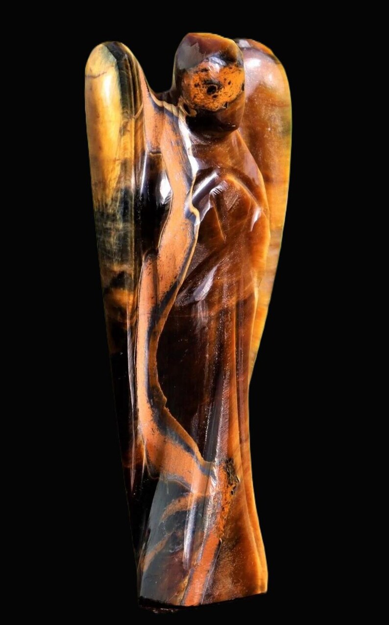 Peut inclure: Une figurine d'ange sculpt&eacute;e en pierre d'&oelig;il de tigre. La sculpture pr&eacute;sente un design d&eacute;taill&eacute; avec un m&eacute;lange de couleurs marron, noir et or. L'ange a des ailes et une expression sereine, sur fond noir.
