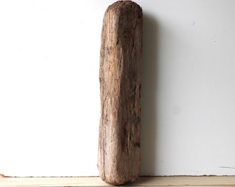 Può includere: Un pezzo di legno rettangolare e invecchiato, con la parte superiore arrotondata. Il legno presenta una texture naturale e ruvida con sfumature di marrone e grigio. È appoggiato contro un muro bianco e una superficie di legno chiaro.