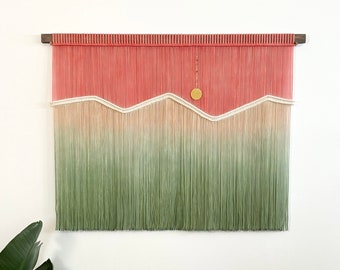 May include: A wall hanging made of yarn with a gradient of pink, beige, and green. The yarn is woven into a pattern of mountains with a small gold circle in the center.