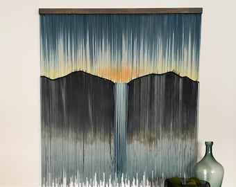 May include: A wall hanging made of yarn with a mountain range design in shades of blue, grey, and yellow. The hanging is mounted on a wooden frame.