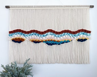May include: A white macrame wall hanging with a woven design featuring blue, red, orange, yellow, and brown yarn. The hanging has a fringe border.