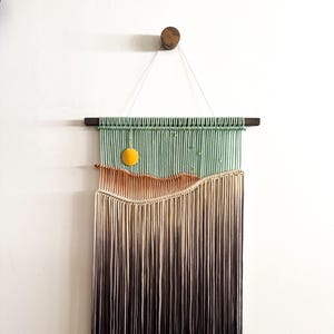 May include: A macrame wall hanging with a green, orange, and brown color gradient. The hanging features a yellow circle in the upper left corner and a wooden dowel at the top.
