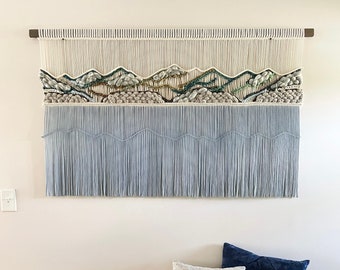 May include: A light blue and white macrame wall hanging with a mountain range design. The wall hanging is hanging on a white wall above a light beige storage bench with two pillows, one gray and one dark blue.