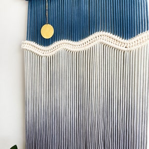 May include: A wall hanging made of macrame with a blue and white ombre design. The macrame is made with thick cotton cord and has a wavy pattern. A gold circle hangs from the top of the macrame.