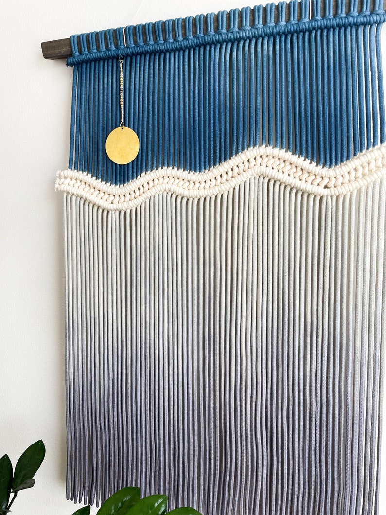 May include: A wall hanging made of macrame with a blue and white ombre design. The macrame is made with thick cotton cord and has a wavy pattern. A gold circle hangs from the top of the macrame.