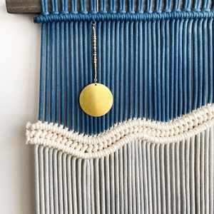 May include: A blue and white macrame wall hanging with a gold circle hanging from the top. The macrame is made with thick cotton cord and has a wave pattern.