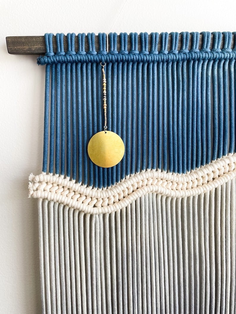 May include: A blue and white macrame wall hanging with a gold circle hanging from the top. The macrame is made with thick cotton cord and has a wave pattern.