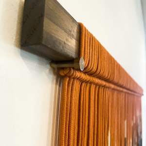 May include: A close-up of a wall hanging made of orange rope, with a wooden dowel at the top. The rope is tied in a pattern that creates a geometric design.