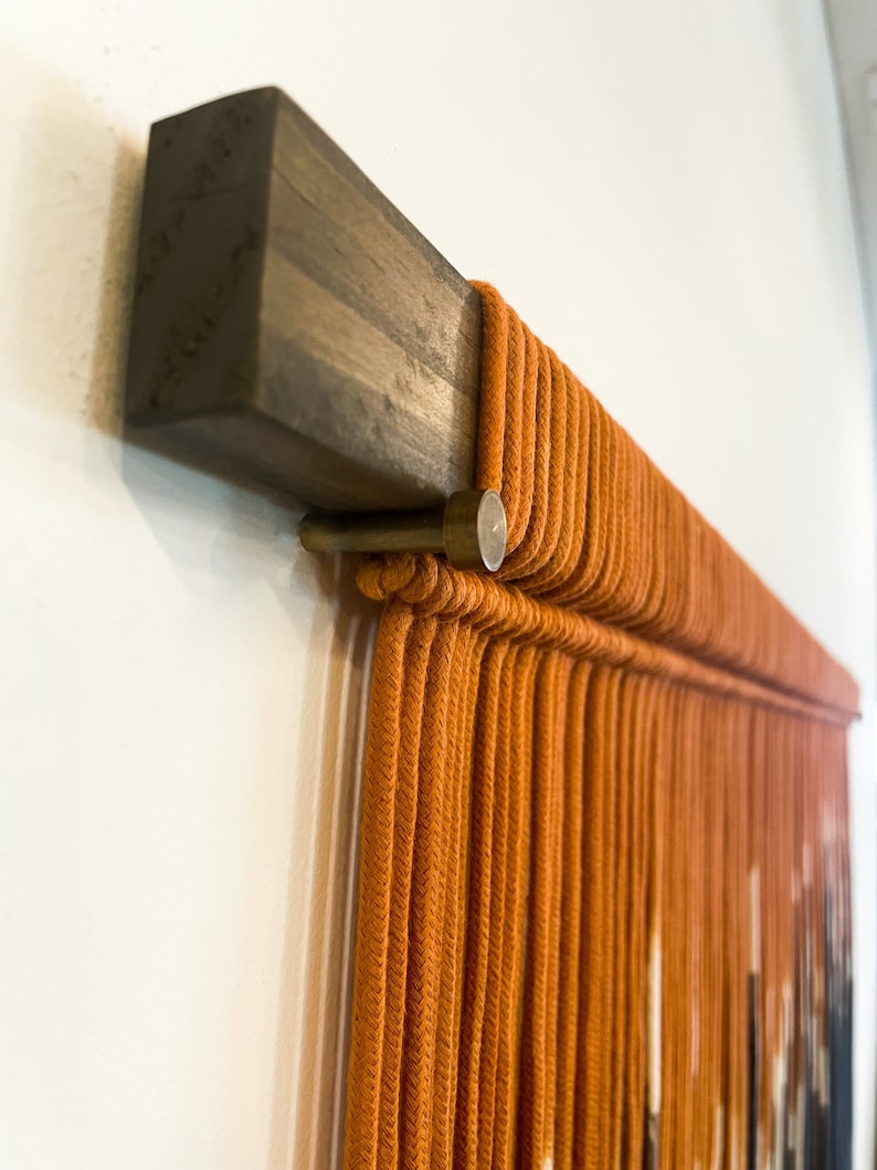 May include: A close-up of a wall hanging made of orange rope, with a wooden dowel at the top. The rope is tied in a pattern that creates a geometric design.
