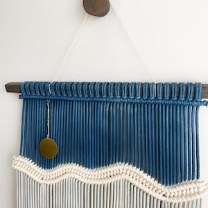 May include: A blue and white macrame wall hanging with a wavy pattern. The hanging features a wooden dowel rod and a brass circle pendant.