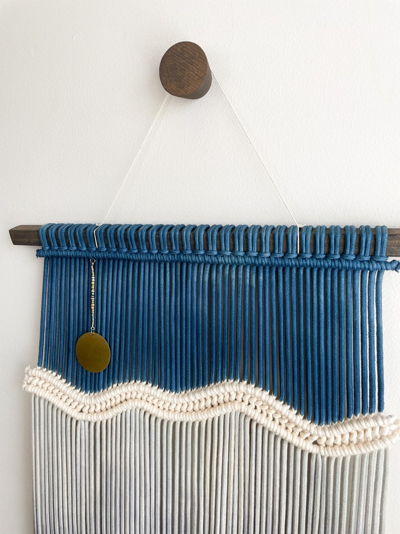 May include: A blue and white macrame wall hanging with a wavy pattern. The hanging features a wooden dowel rod and a brass circle pendant.