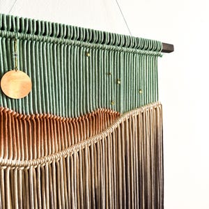 May include: A green, brown, and beige macrame wall hanging with gold beads and a wooden circle. The hanging is attached to a wooden dowel rod.