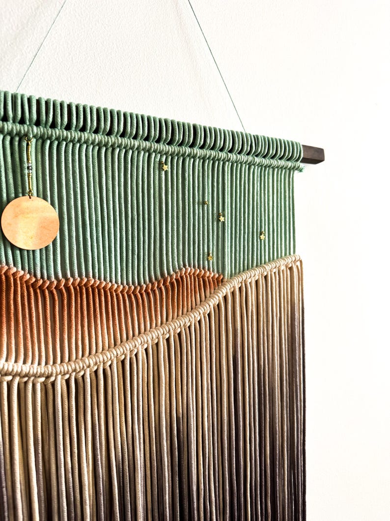 May include: A green, brown, and beige macrame wall hanging with gold beads and a wooden circle. The hanging is attached to a wooden dowel rod.