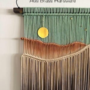 May include: A macrame wall hanging with a green, orange, and brown color gradient. The hanging features a brass circle and gold stars. The text "Add Brass Hardware" is written on the top of the image.