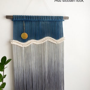 May include: A blue and white ombre macrame wall hanging with a wooden dowel at the top and a small yellow circle hanging from the top. The wall hanging is attached to a white wall with a wooden hook.