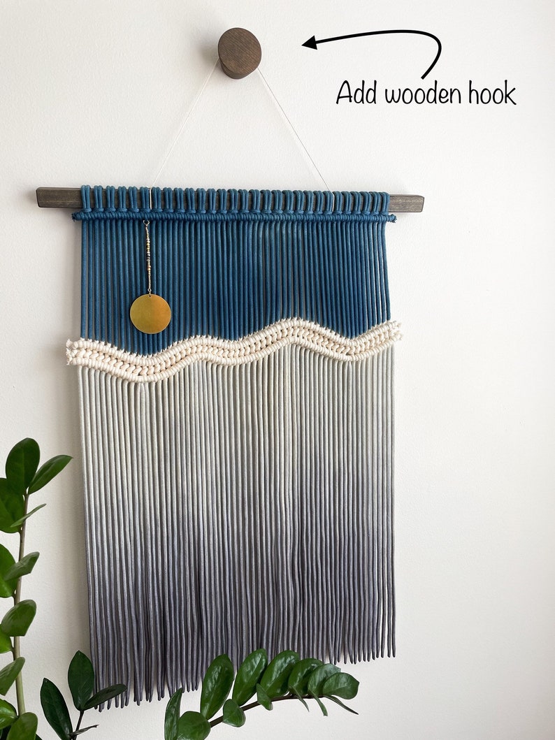 May include: A blue and white ombre macrame wall hanging with a wooden dowel at the top and a small yellow circle hanging from the top. The wall hanging is attached to a white wall with a wooden hook.