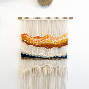 May include: A white macrame wall hanging with a woven design featuring orange, yellow, brown, blue, and cream colored yarn. The hanging has a fringe at the bottom.