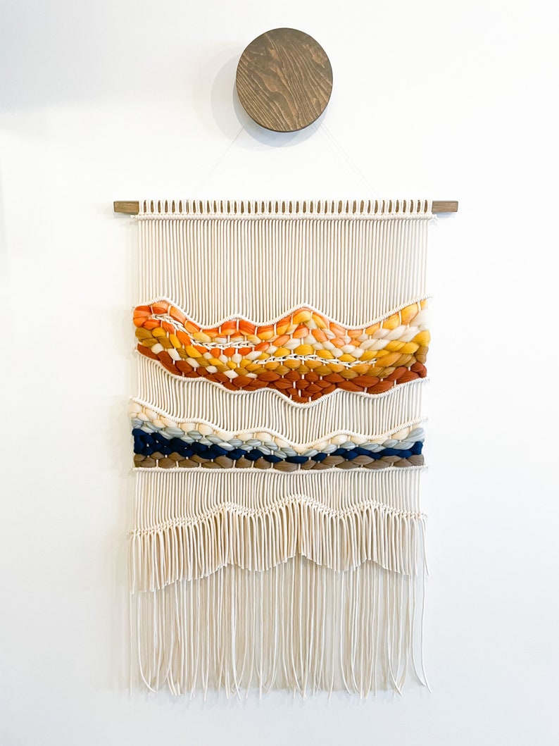 May include: A white macrame wall hanging with a woven design featuring orange, yellow, brown, blue, and cream colored yarn. The hanging has a fringe at the bottom.