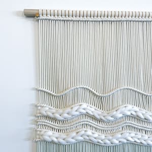 May include: White and light blue macrame wall hanging with a woven pattern. The hanging is made with cotton cord and has a wooden dowel at the top.