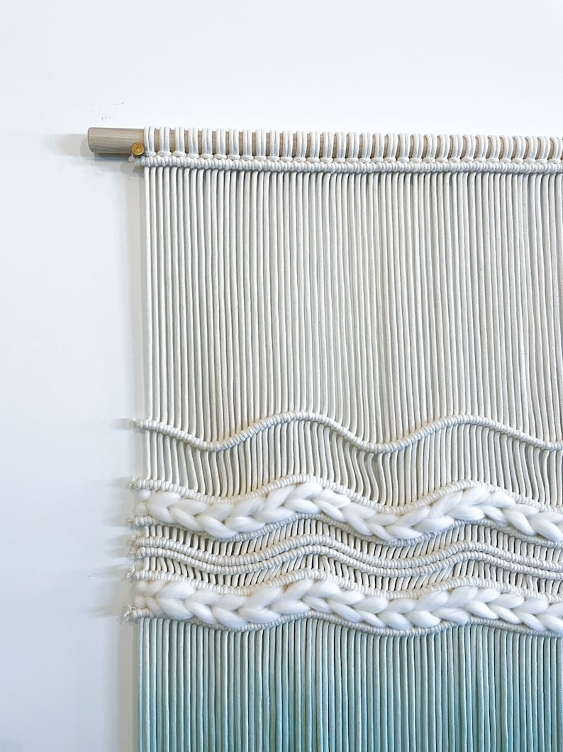 May include: White and light blue macrame wall hanging with a woven pattern. The hanging is made with cotton cord and has a wooden dowel at the top.