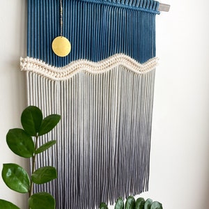 May include: A blue and white macrame wall hanging with a wavy design and a gold circle ornament. The hanging is made with cotton cord and has a wooden dowel at the top.