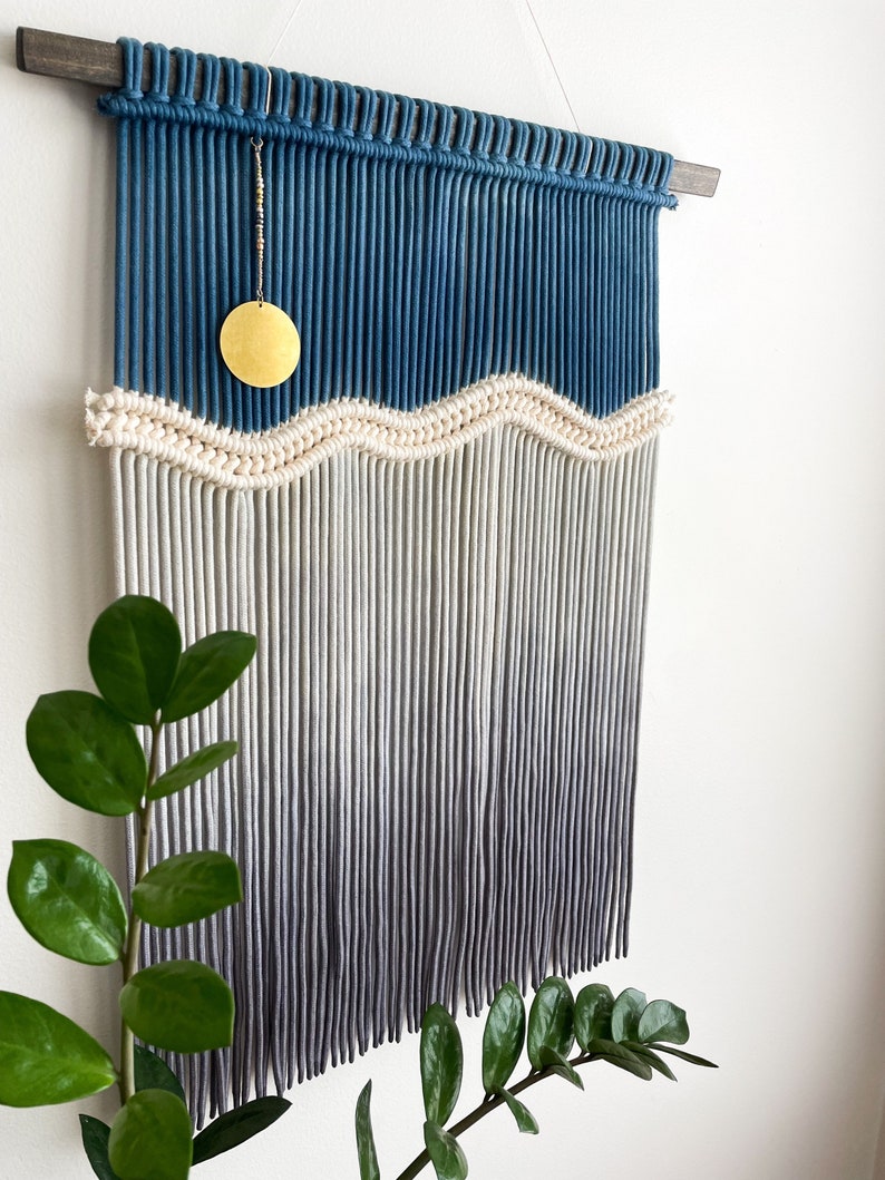 May include: A blue and white macrame wall hanging with a wavy design and a gold circle ornament. The hanging is made with cotton cord and has a wooden dowel at the top.