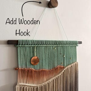 May include: A macrame wall hanging with a green, orange, and brown color gradient. The hanging is attached to a wooden dowel rod and a wooden hook. A copper circle hangs from the top of the hanging.