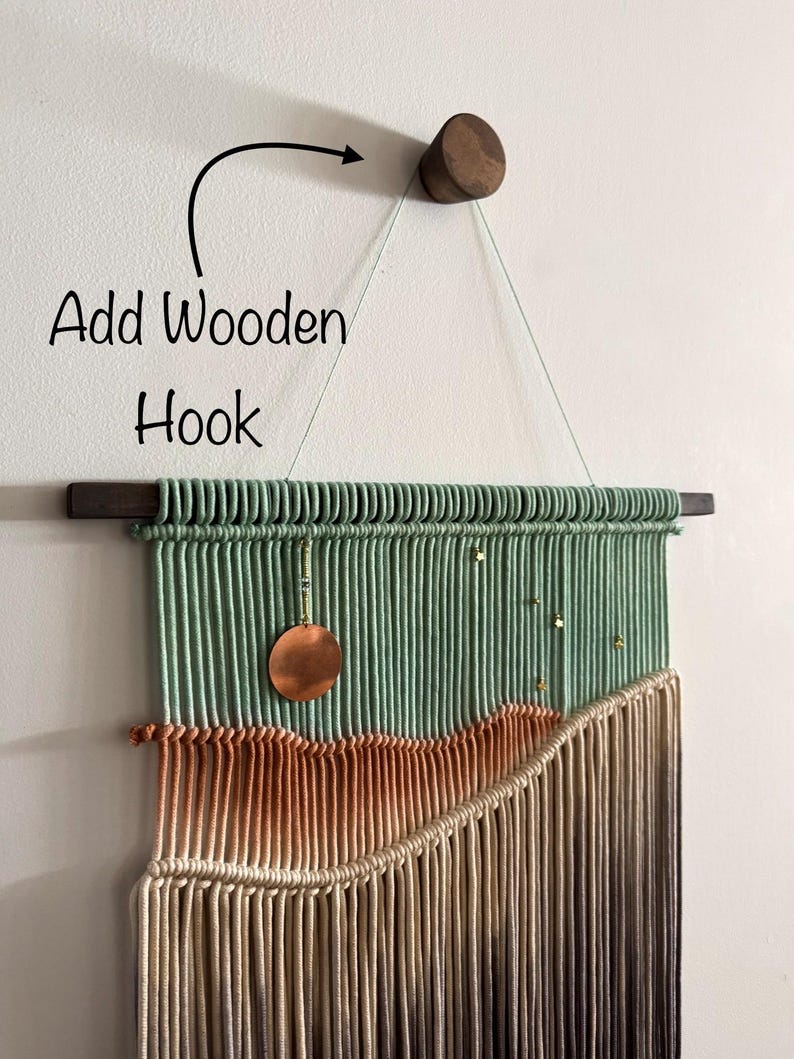 May include: A macrame wall hanging with a green, orange, and brown color gradient. The hanging is attached to a wooden dowel rod and a wooden hook. A copper circle hangs from the top of the hanging.