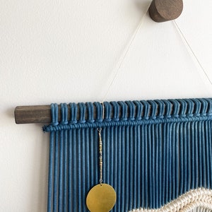 May include: A blue and white macrame wall hanging with a gold circle pendant. The hanging is attached to a dark wood dowel rod and a dark wood wall hook.