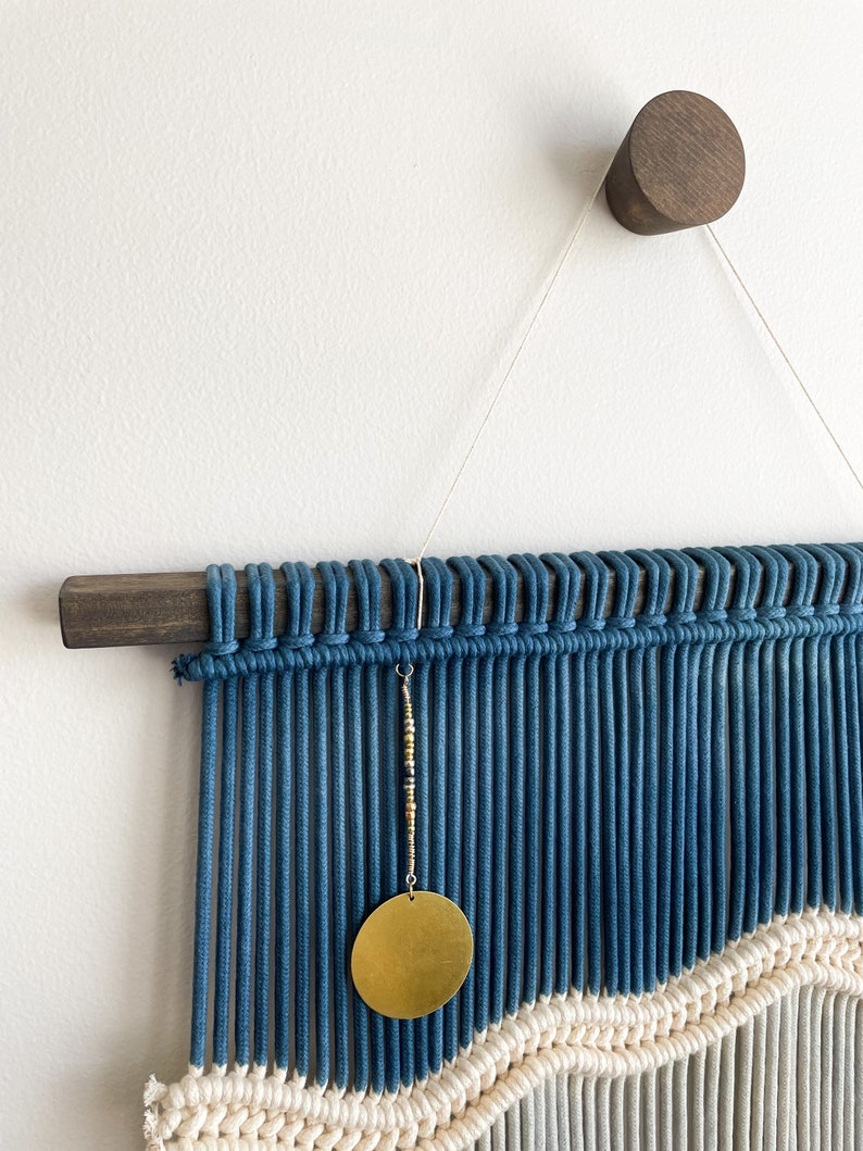 May include: A blue and white macrame wall hanging with a gold circle pendant. The hanging is attached to a dark wood dowel rod and a dark wood wall hook.