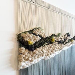 May include: A wall hanging made of macrame with a textured design featuring a variety of colors, including white, green, and yellow. The hanging has a fringe of blue yarn.
