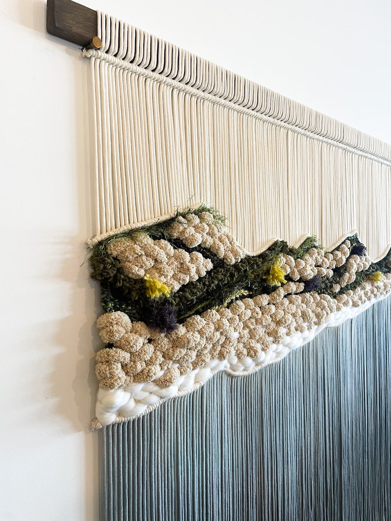 May include: A wall hanging made of macrame with a textured design featuring a variety of colors, including white, green, and yellow. The hanging has a fringe of blue yarn.