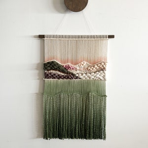 以下が含まれることがあります： 緑、ピンク、白の織り模様が施された壁掛け。下部に緑色の糸のフリンジが付いている。