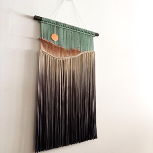 May include: A wall hanging made of yarn with a green, orange, and brown color gradient. The yarn is woven into a pattern that resembles a landscape. The hanging is attached to a wooden dowel rod.