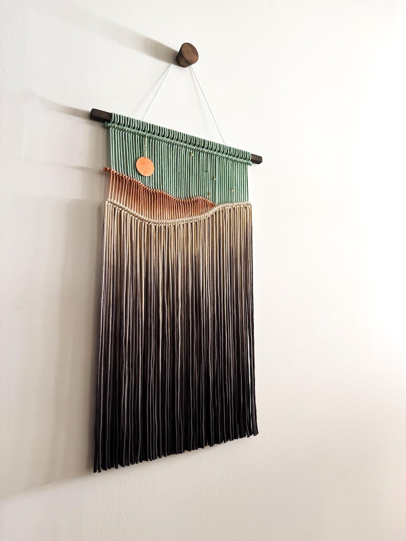 May include: A wall hanging made of yarn with a green, orange, and brown color gradient. The yarn is woven into a pattern that resembles a landscape. The hanging is attached to a wooden dowel rod.