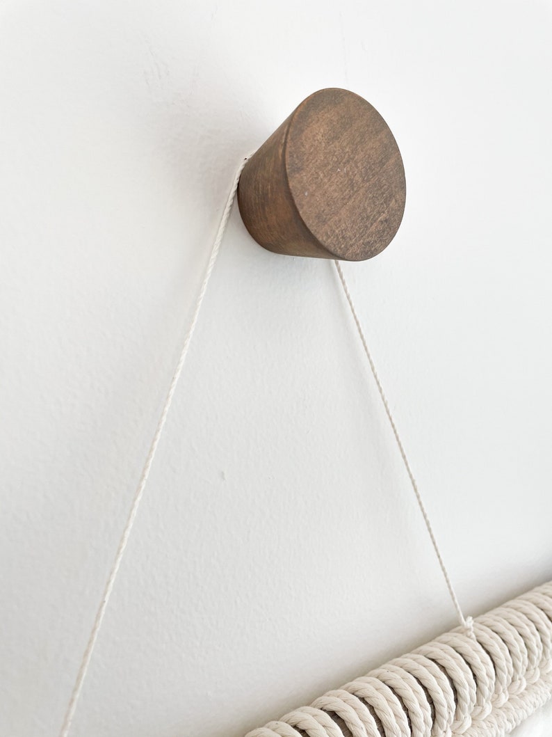 May include: A wooden wall hook with a rounded top, attached to a white wall with a white rope hanging from it.