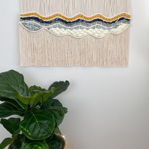 May include: A macrame wall hanging with a wavy pattern in shades of white, gray, and yellow. The hanging has a fringe border and is mounted on a wooden dowel.