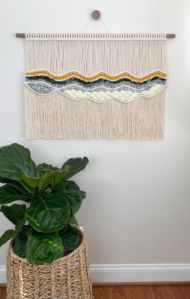 May include: A macrame wall hanging with a wavy pattern in shades of white, gray, and yellow. The hanging has a fringe border and is mounted on a wooden dowel.