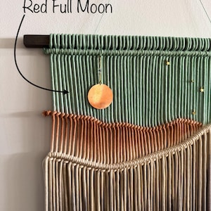 May include: A green, orange, and beige macrame wall hanging with a copper moon and gold stars. The hanging is labeled "Red Full Moon".