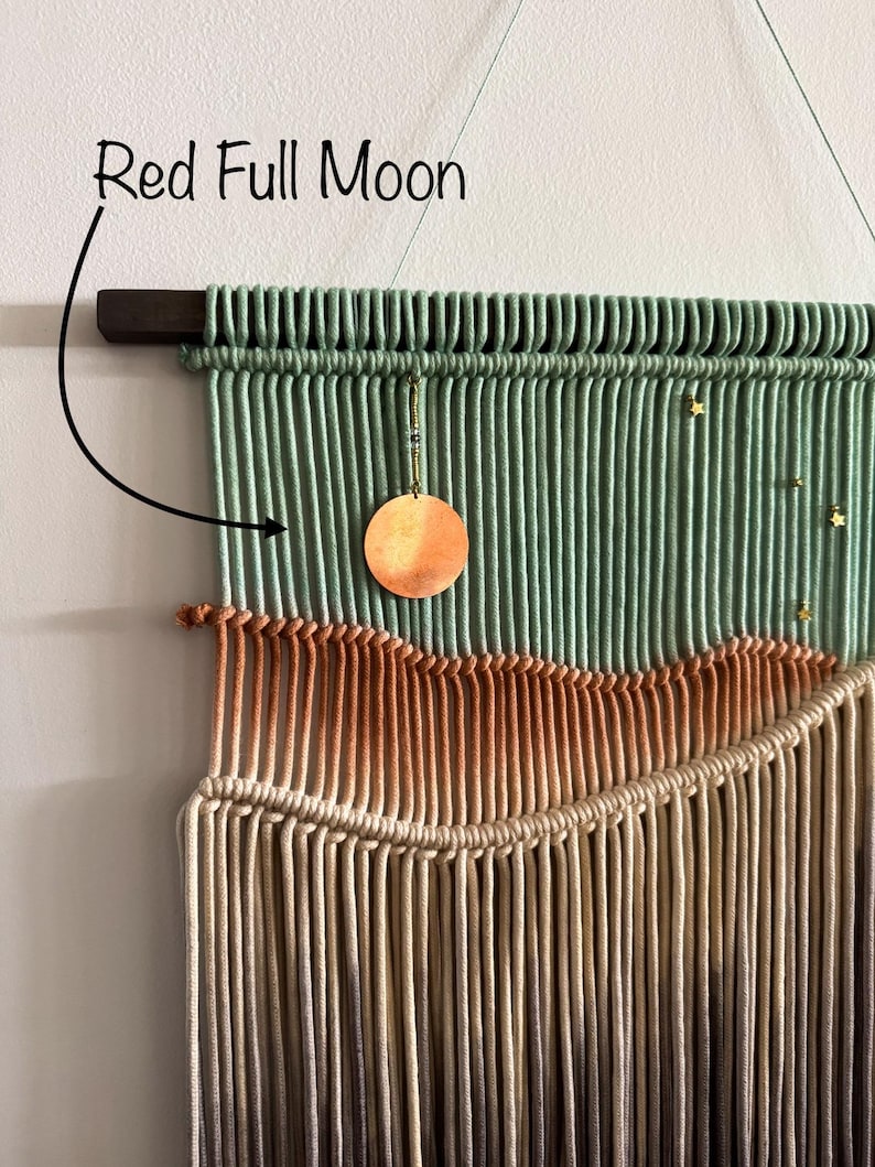 May include: A green, orange, and beige macrame wall hanging with a copper moon and gold stars. The hanging is labeled "Red Full Moon".