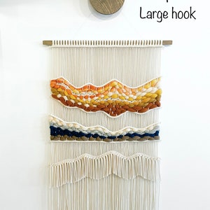May include: A white macrame wall hanging with a woven design featuring orange, yellow, brown, blue, and white yarn. The hanging has a fringe at the bottom and is hung on a wooden dowel rod.