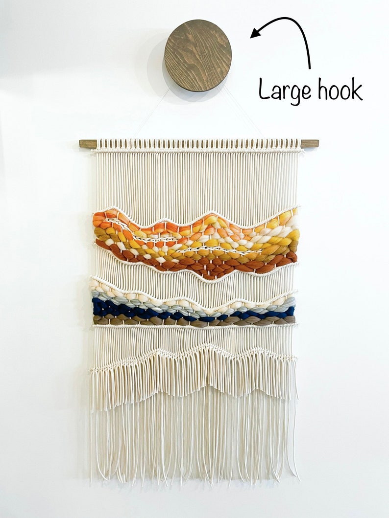 May include: A white macrame wall hanging with a woven design featuring orange, yellow, brown, blue, and white yarn. The hanging has a fringe at the bottom and is hung on a wooden dowel rod.