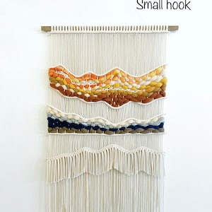 May include: A white macrame wall hanging with a woven design in shades of orange, yellow, blue, and brown. The hanging has a wooden dowel at the top and a small hook for hanging.