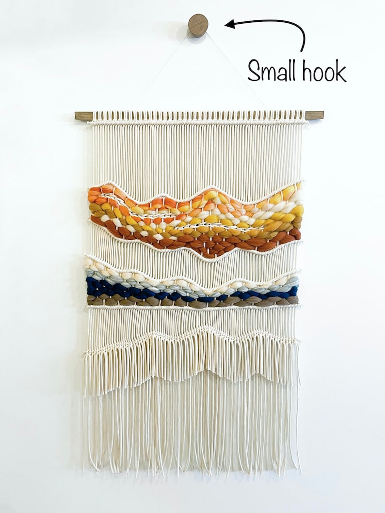 May include: A white macrame wall hanging with a woven design in shades of orange, yellow, blue, and brown. The hanging has a wooden dowel at the top and a small hook for hanging.