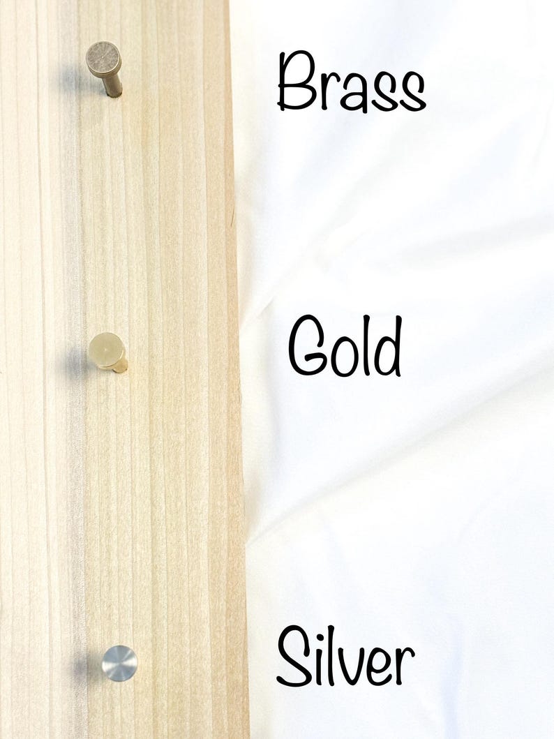 May include: Three metal hooks in brass, gold, and silver finishes mounted on a wooden board.
