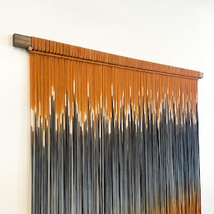 May include: A wall hanging made of macrame with a gradient of orange, white, and blue yarn. The yarn is tied together in a series of knots to create a textured and visually appealing design. The wall hanging is attached to a wooden dowel.