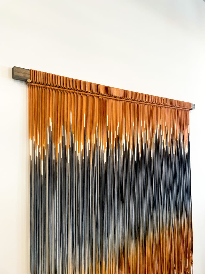 May include: A wall hanging made of macrame with a gradient of orange, white, and blue yarn. The yarn is tied together in a series of knots to create a textured and visually appealing design. The wall hanging is attached to a wooden dowel.